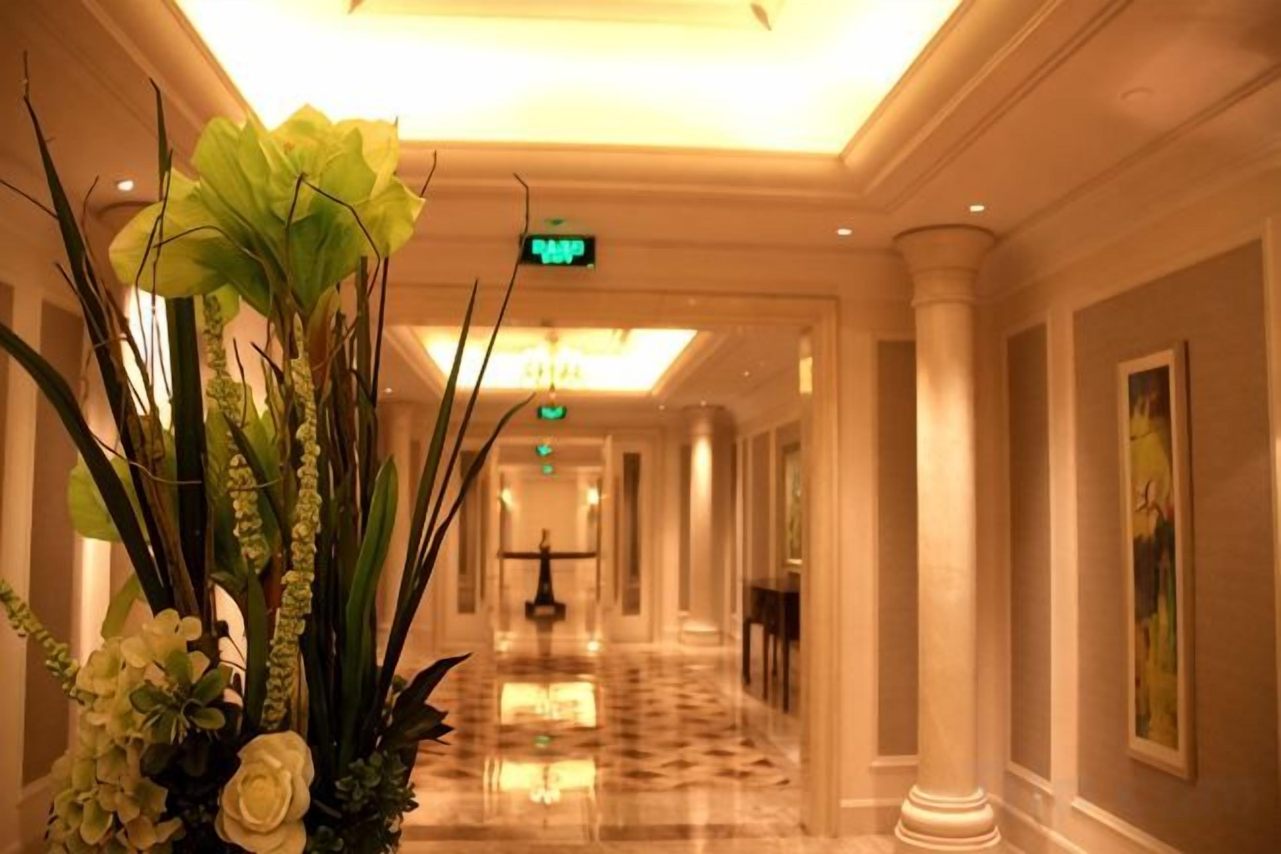 Un pasillo de hotel con piso de mármol pulido, un letrero de salida verde y un gran arreglo floral.
