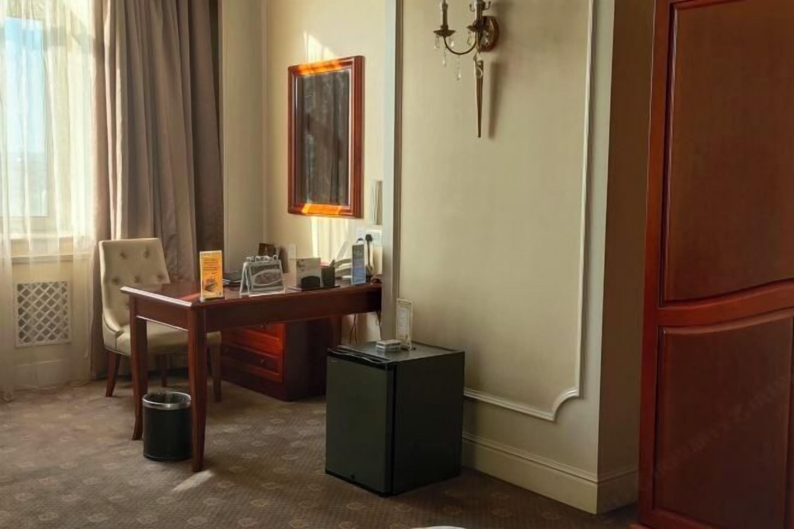 Et hotelværelse med skrivebord, stol, lille køleskab og vægmonteret lampe.