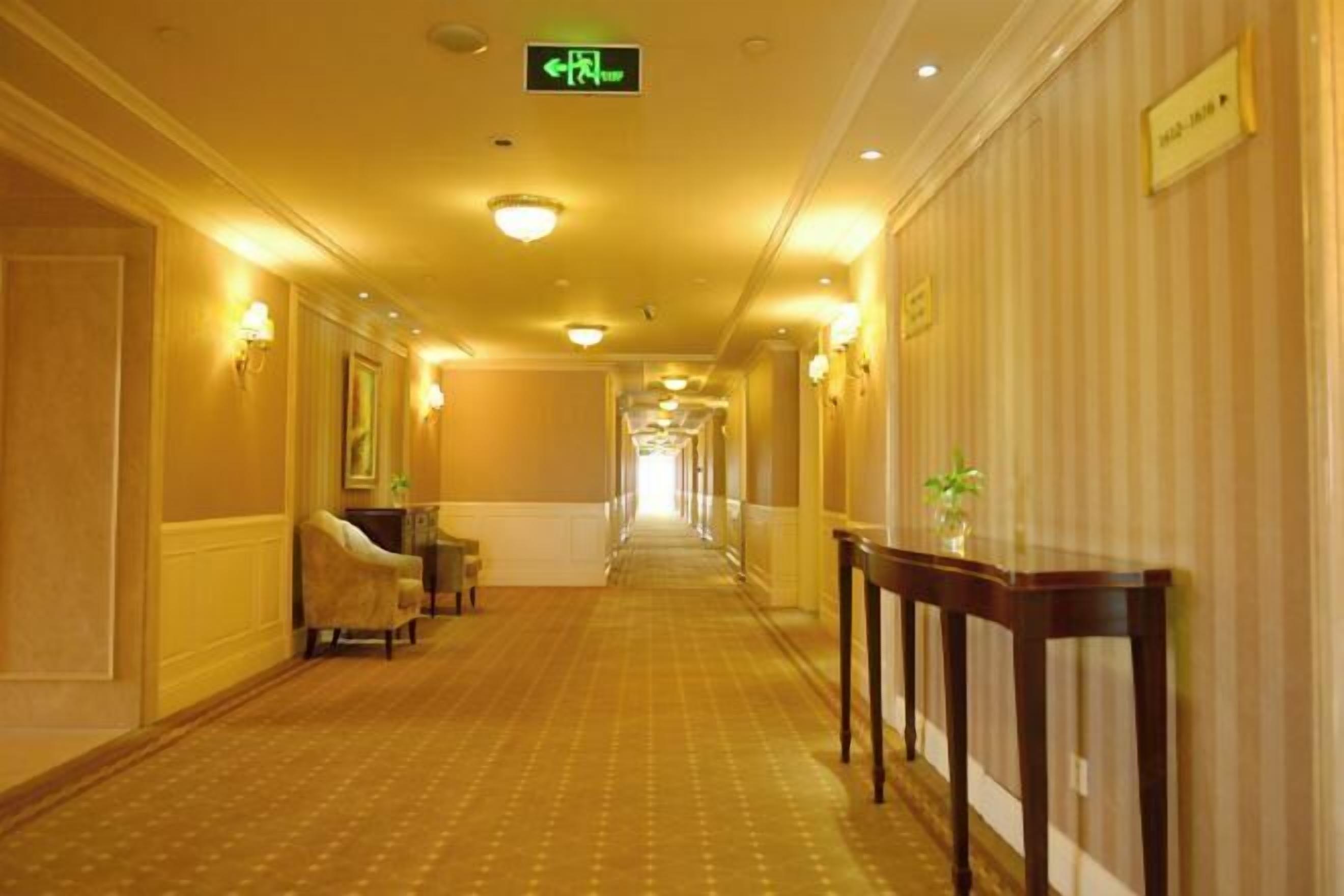 En hotellkorridor med ett heltäckningsmatta på golvet, väggmonterade lampor och en skylt som visar vägen till utgången.