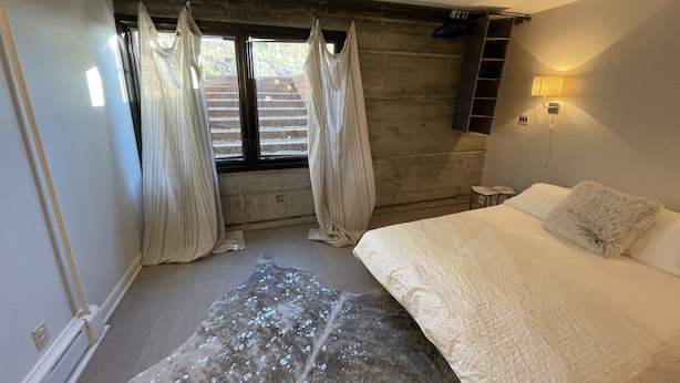 A bedroom with a bed, a window with curtains, a rug, and a wall-mounted shelf.