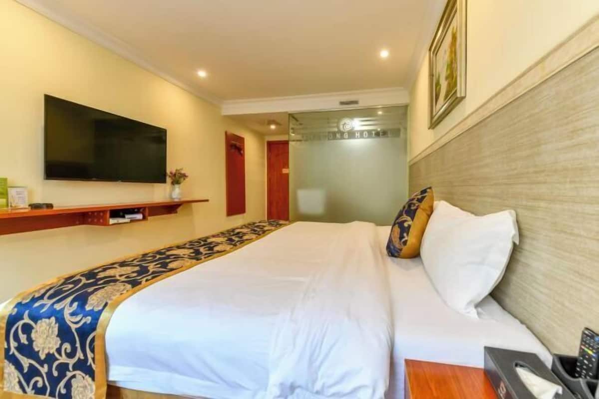 Una habitación de hotel con una cama, un televisor montado en la pared, una mesita de noche con un teléfono y una caja de pañuelos, y una puerta que da a otra habitación.