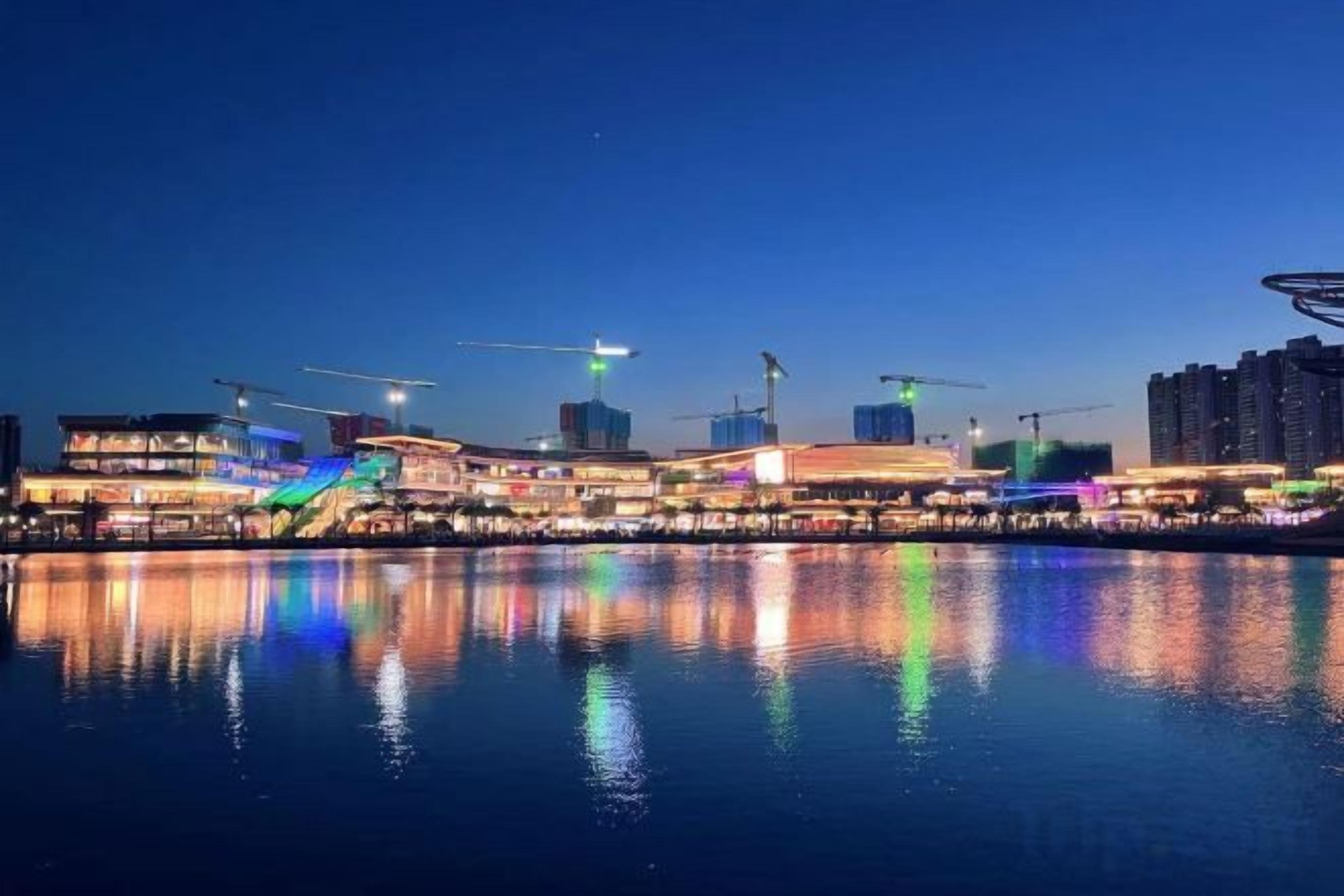 A waterfront cityscape at night with illuminated buildings and a calm river reflecting the lights.