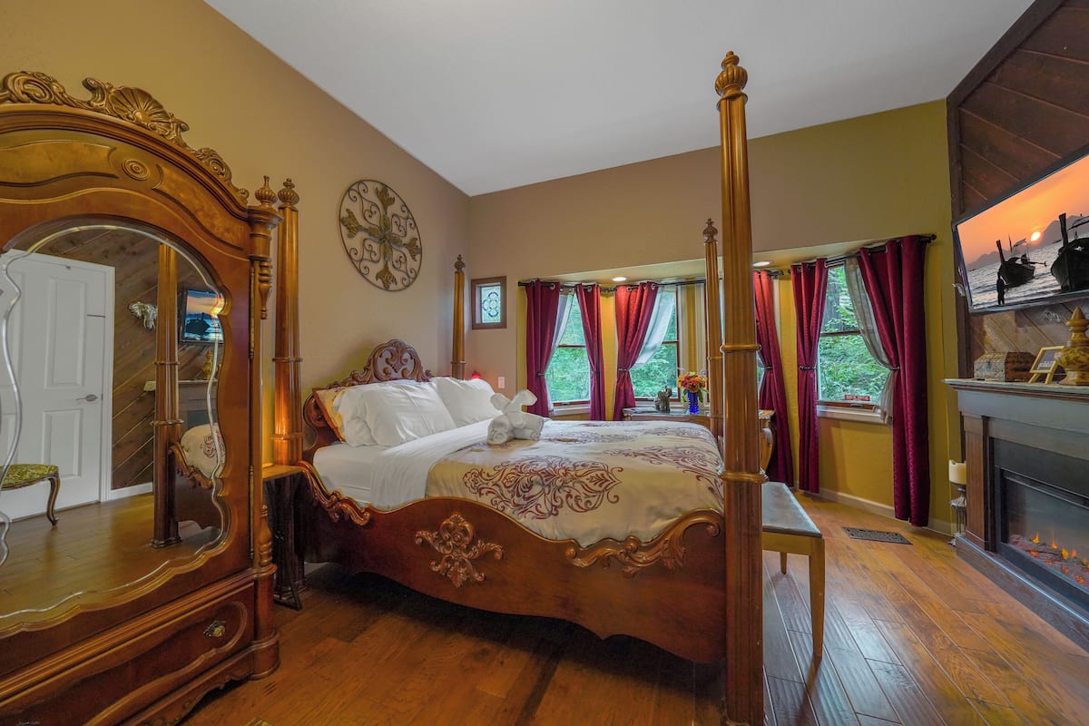 A four-poster bed with a mirror, a fireplace, and a view of the sunset through windows.