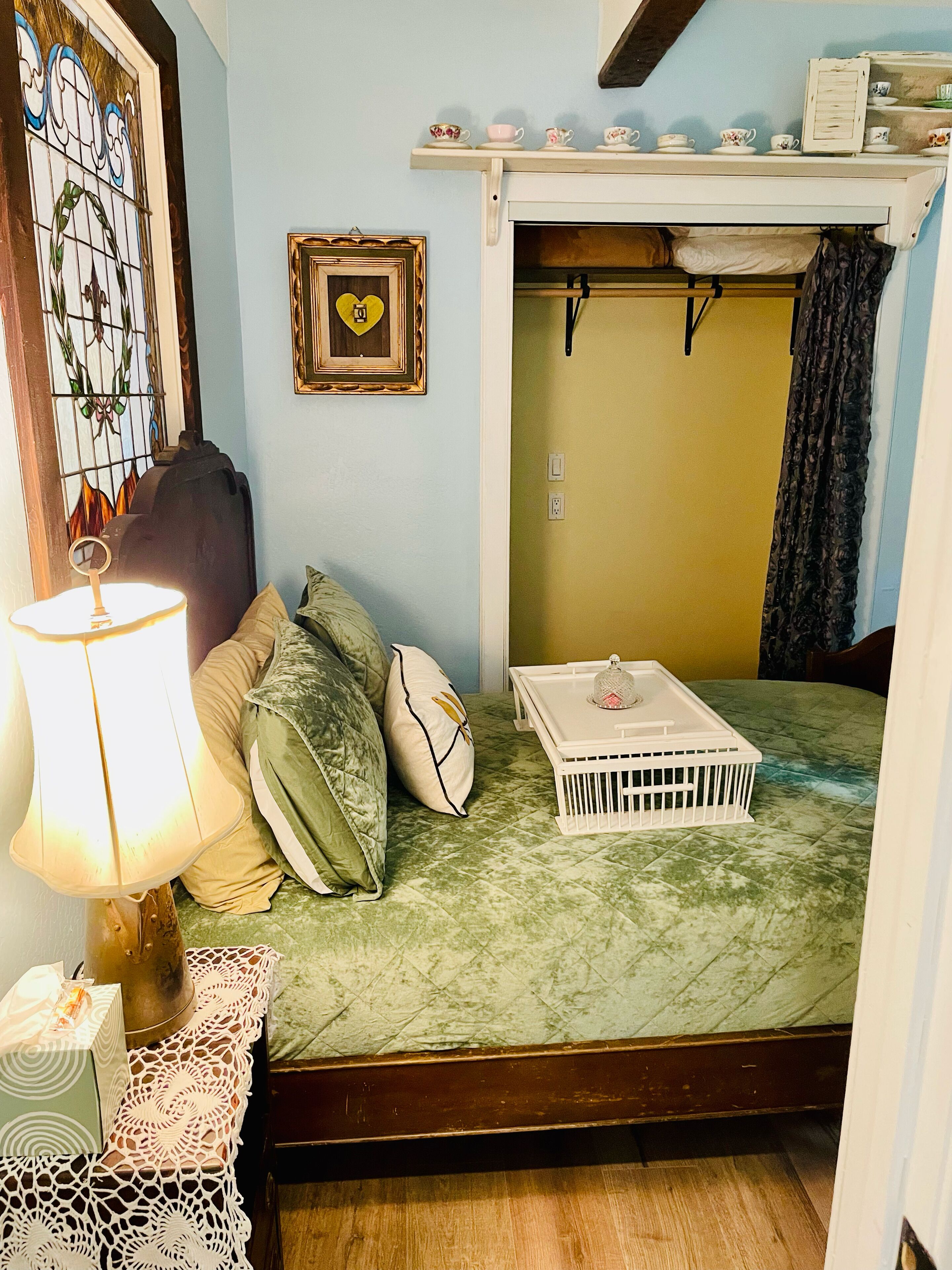 A small, cozy room with a bed, a bedside table with a lamp, a suitcase, and a wall-mounted shelf with decorative items.
