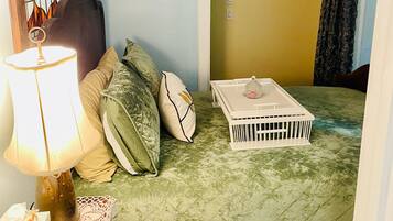 A small, cozy room with a bed, a bedside table with a lamp, a suitcase, and a wall-mounted shelf with decorative items.