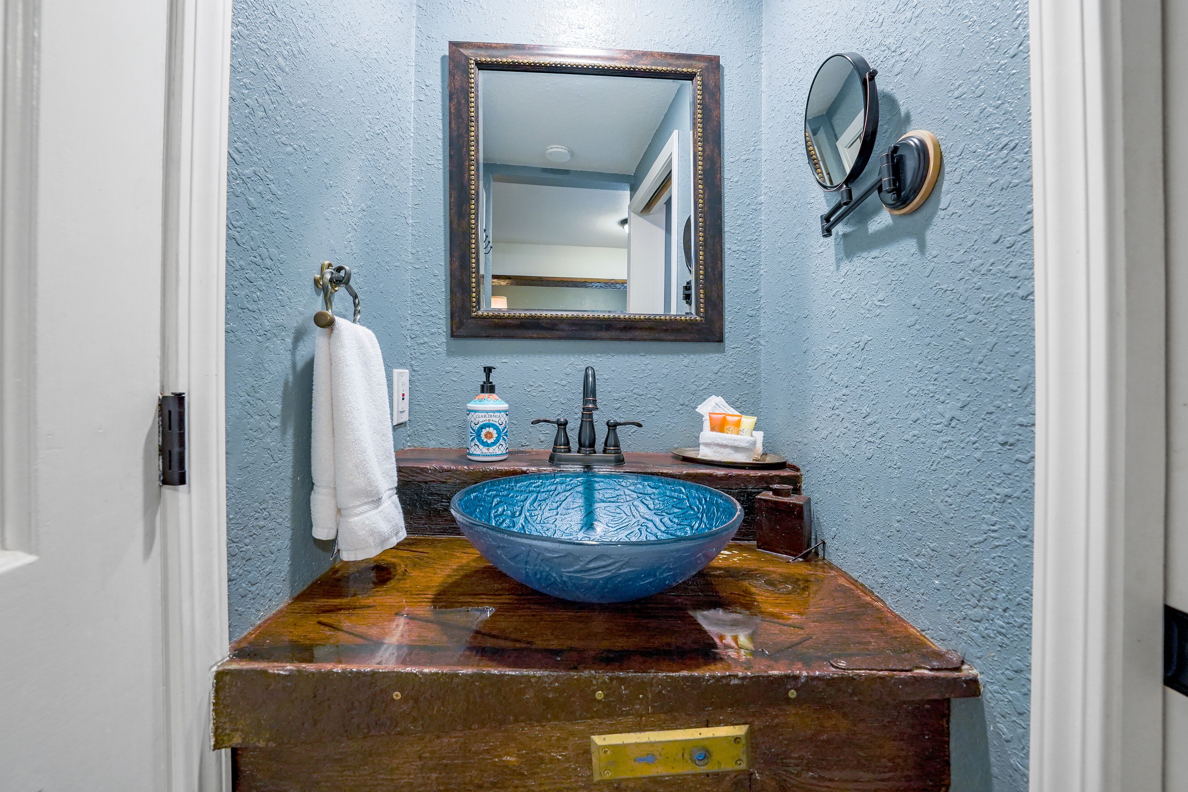 Un baño con un lavabo de vidrio azul, un tocador de madera, una toalla blanca colgada en un perchero y un espejo con marco de madera, además de una lámpara de pared.