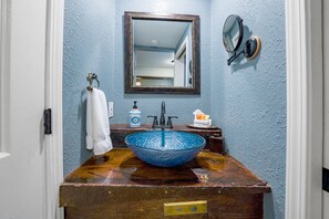 A bathroom with a blue glass sink, a wooden vanity, a white towel hanging on a towel rack, a mirror with a wooden frame, and a wall-mounted light fixture. - Flume's End (Nevada City)