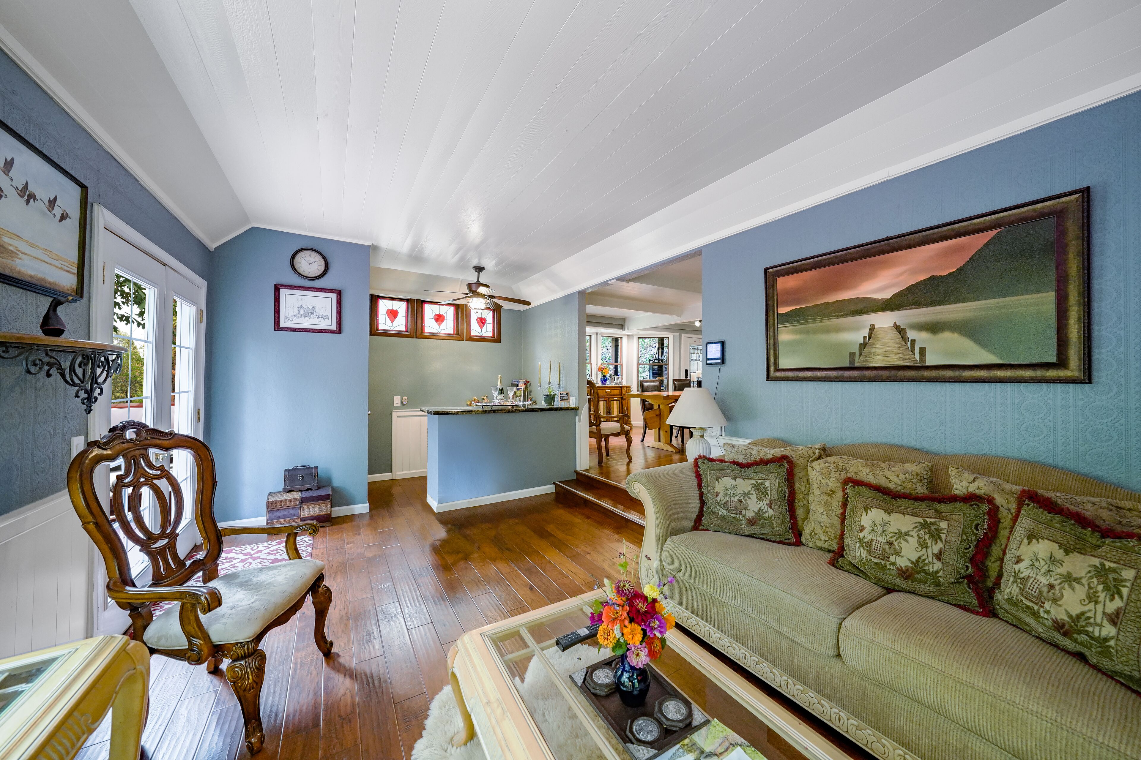 A spacious living room with a wooden floor, a green sofa with patterned cushions, a wooden chair, and a coffee table with a vase of flowers.