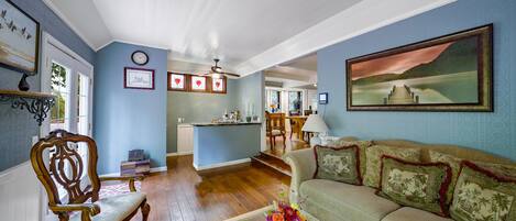 A spacious living room with a wooden floor, a green sofa with patterned cushions, a wooden chair, and a coffee table with a vase of flowers.