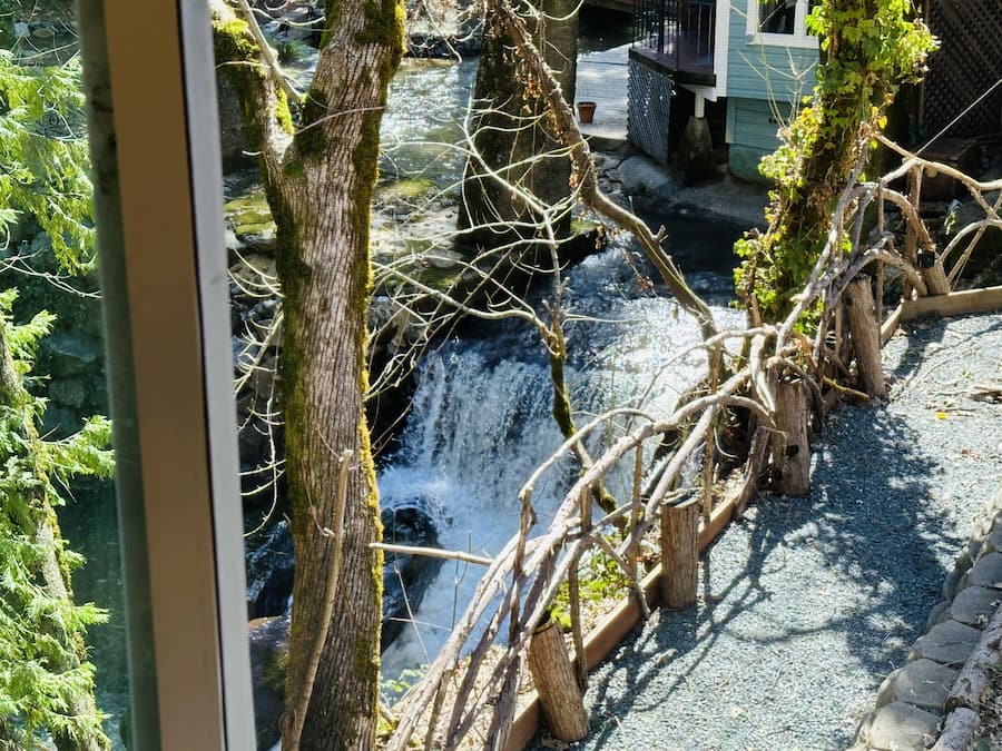 A view of a stone bridge over a small waterfall, surrounded by trees and greenery.