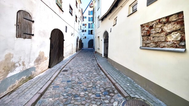 Une ruelle étroite pavée, bordée de bâtiments historiques dotés d’entrées en arc de cercle et de volets traditionnels.