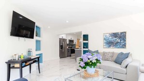 A modern living room with a flat-screen TV, a sofa, a coffee table, and a kitchen in the background. - WPB Elegant 5BR | Hot Tub | Fire Pit | Near Beach (West Palm Beach)