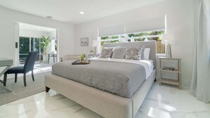 A modern bedroom with a large bed, a chair, a small table, and a window with blinds. - WPB Elegant 5BR | Hot Tub | Fire Pit | Near Beach (West Palm Beach)