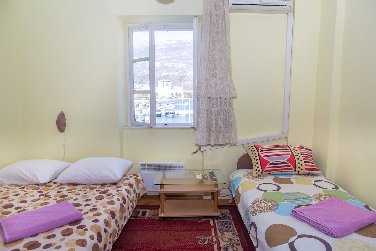 A small, well-lit room with two beds, a window with a view of boats and mountains, and a red rug.