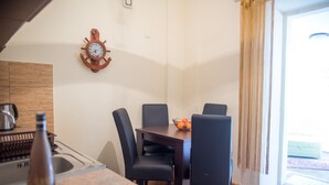 A dining area with a wooden table, four chairs, a clock, and a bowl of fruit on the table. - Apartments Old Town Aleksici (Budva)