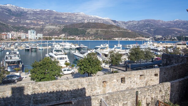 A marina with boats docked, a stone wall in the foreground, and mountains in the background. - Apartments Old Town Aleksici (Budva)