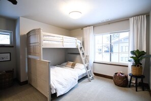 A bunk bed room with a ladder, a window with curtains, a plant on a side table, and a basket on the floor.