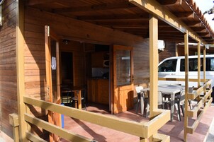 Una cabaña de madera con un porche cubierto, una zona de cocina y una camioneta blanca estacionada afuera.