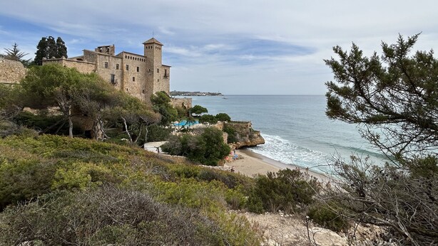 A historic stone castle with a tower, overlooking a beach with a swimming pool and rocky coastline.