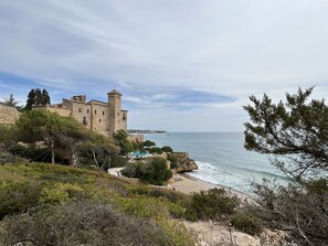 Un château historique en pierre, doté d’une tour, qui surplombe une plage avec une piscine et une côte rocheuse.