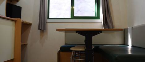 Una habitación pequeña con una mesa, un banco, un taburete, un televisor y un ventanal con cortinas.