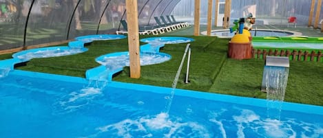 Zona de piscina exterior cubierta con fuente de agua, terraza de madera y área de juegos cercada.