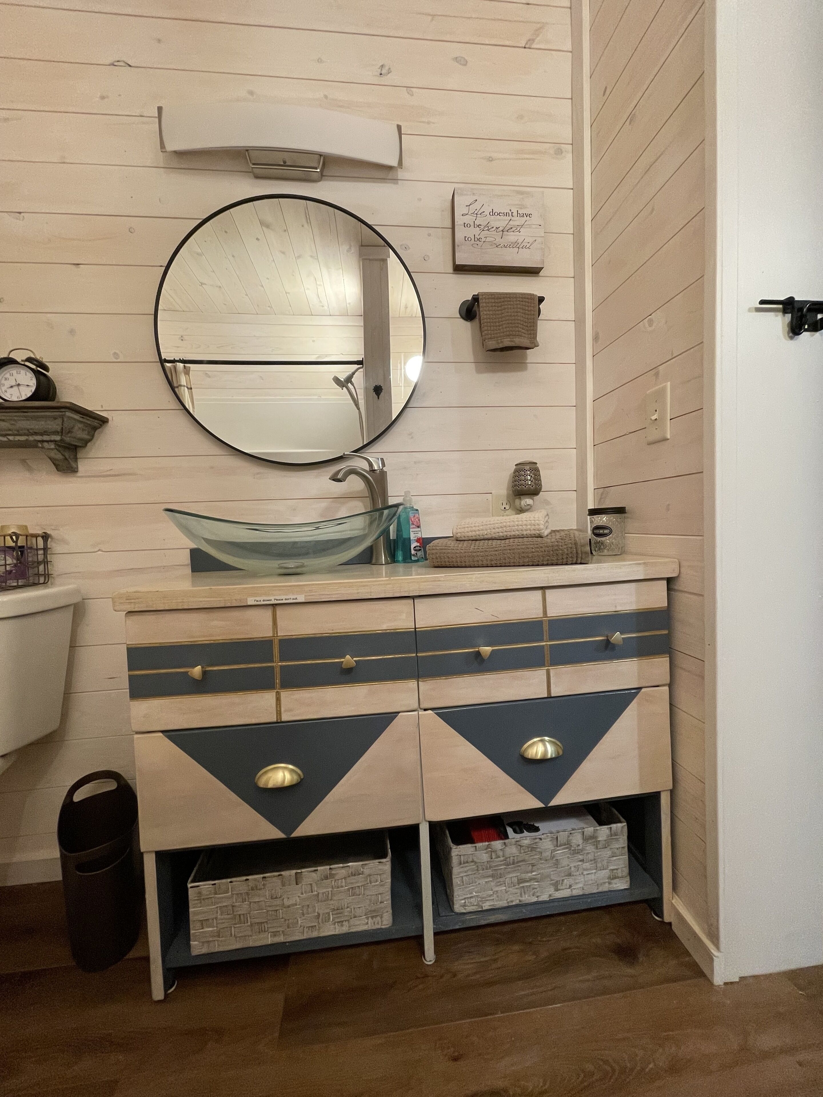 Une salle de bain avec un miroir rond, un lavabo en verre et un meuble-lavabo doté de tiroirs et d’une poubelle.