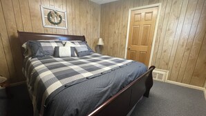 A bedroom with a wooden paneled wall, a bed with a checkered bedspread, a nightstand, a lamp, and a door. - All-inclusive Apartment - 1 Bedroom, 1 Bath, Equipped Kitchen, & Laundry (Terre Haute)