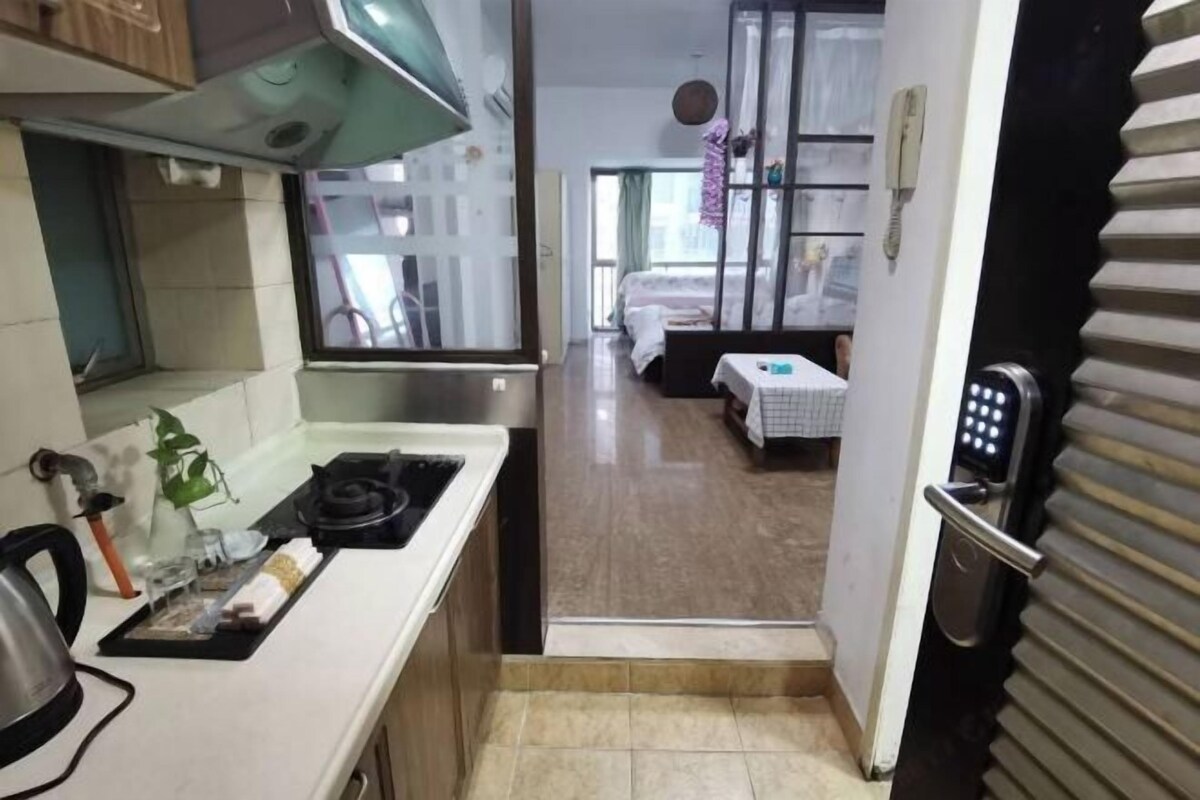 A narrow kitchen with a gas stove, a kettle, and a small countertop. A door leads to a bedroom with a bed and a nightstand.