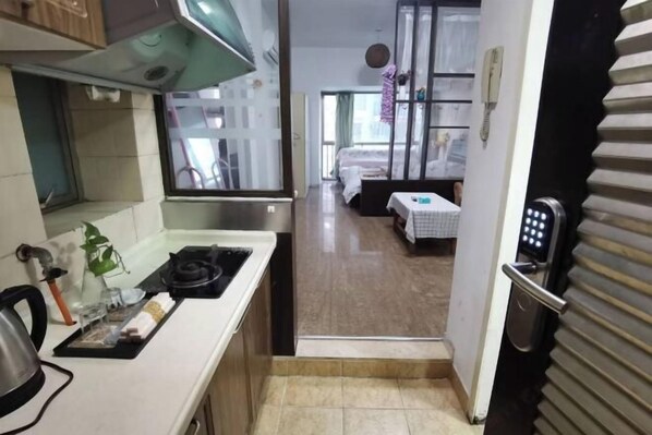 A narrow kitchen with a gas stove, a kettle, and a small countertop. A door leads to a bedroom with a bed and a nightstand. - Shenzhen Four Seasons Family Apartment (Shenzhen)