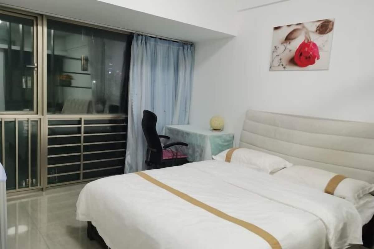A hotel room with a bed, a small table, a chair, a window with curtains, and a balcony.