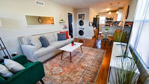 A modern living room with a sofa, a green armchair, a coffee table, and a patterned rug. There is a kitchen area with a dining table and chairs, and a laundry area with a washing machine and dryer. - ★ Cozy Escape★ Central Home ★ Plaza KC (Kansas City)
