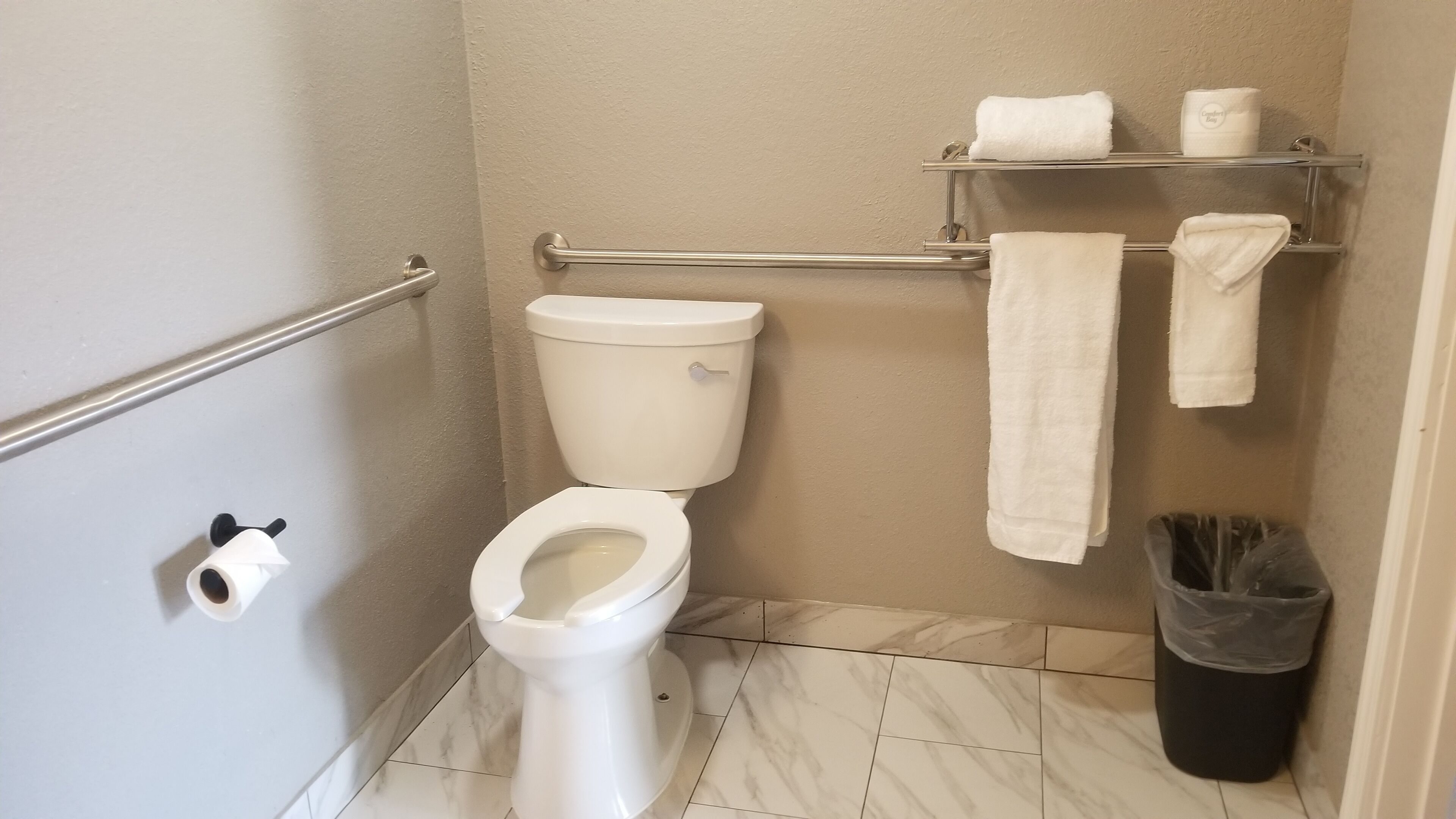 A small bathroom with a toilet, a handrail, a towel rack with towels, a trash bin, and a toilet paper holder.