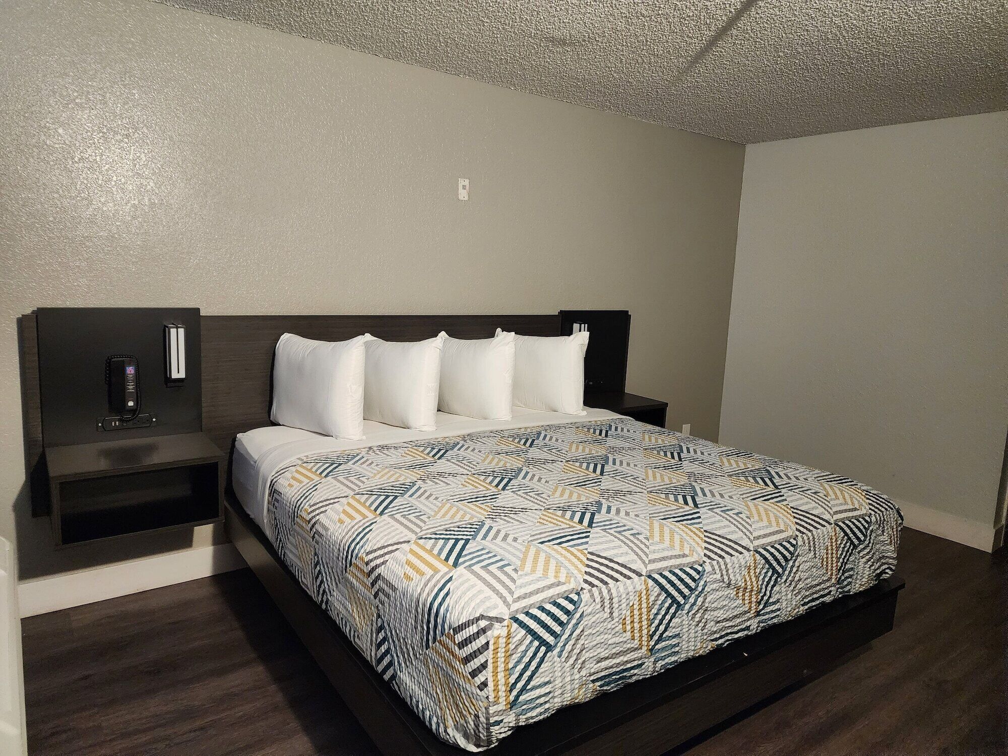 A neatly made bed with a patterned bedspread, a bedside table with a phone, and a wall-mounted light fixture.