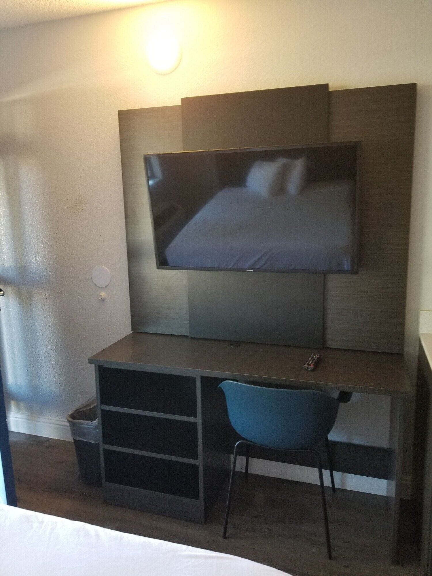 A hotel room with a bed, a desk with a chair, a television mounted on the wall, and a trash bin.