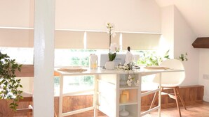 A bright, spacious room with a high ceiling and wooden beams, featuring a white table with a vase of flowers, a chair, and a window with a view of greenery.