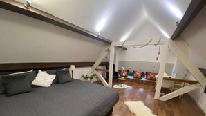 A bedroom with a large bed, wooden flooring, a white fur rug, and a view of a dining area with a staircase.