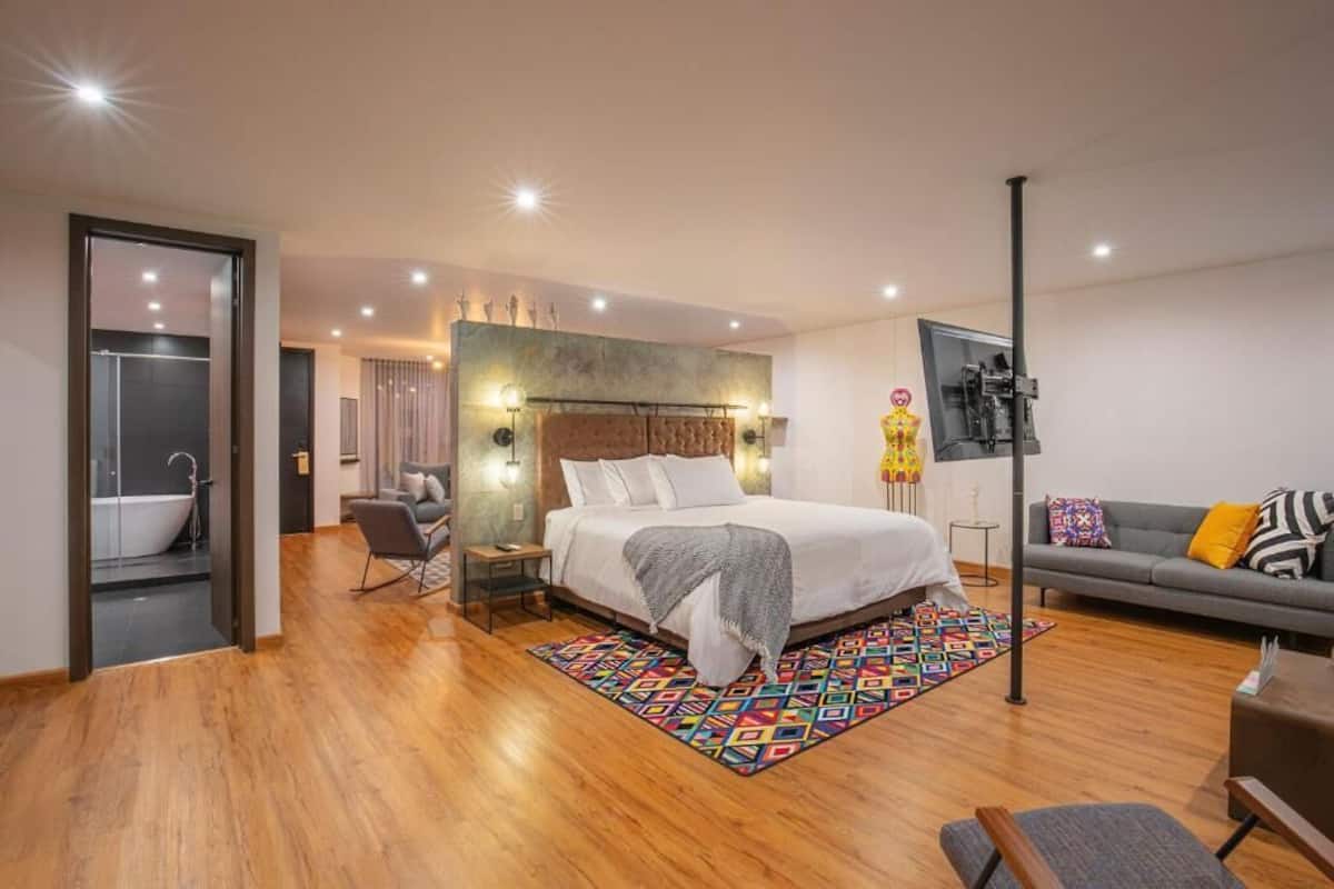 Une chambre moderne avec un grand lit, un coin salon avec un canapé, une petite table et un tapis coloré.