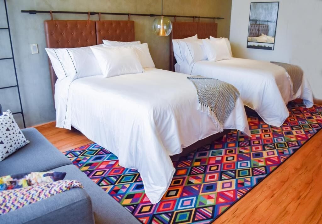 A hotel room with two beds, a gray sofa, and a colorful geometric-patterned rug.