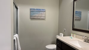 A bathroom with a white toilet, a sink with a mirror, and a painting of a beach scene on the wall. - Aspire Inn & Suites Orange (Orange)