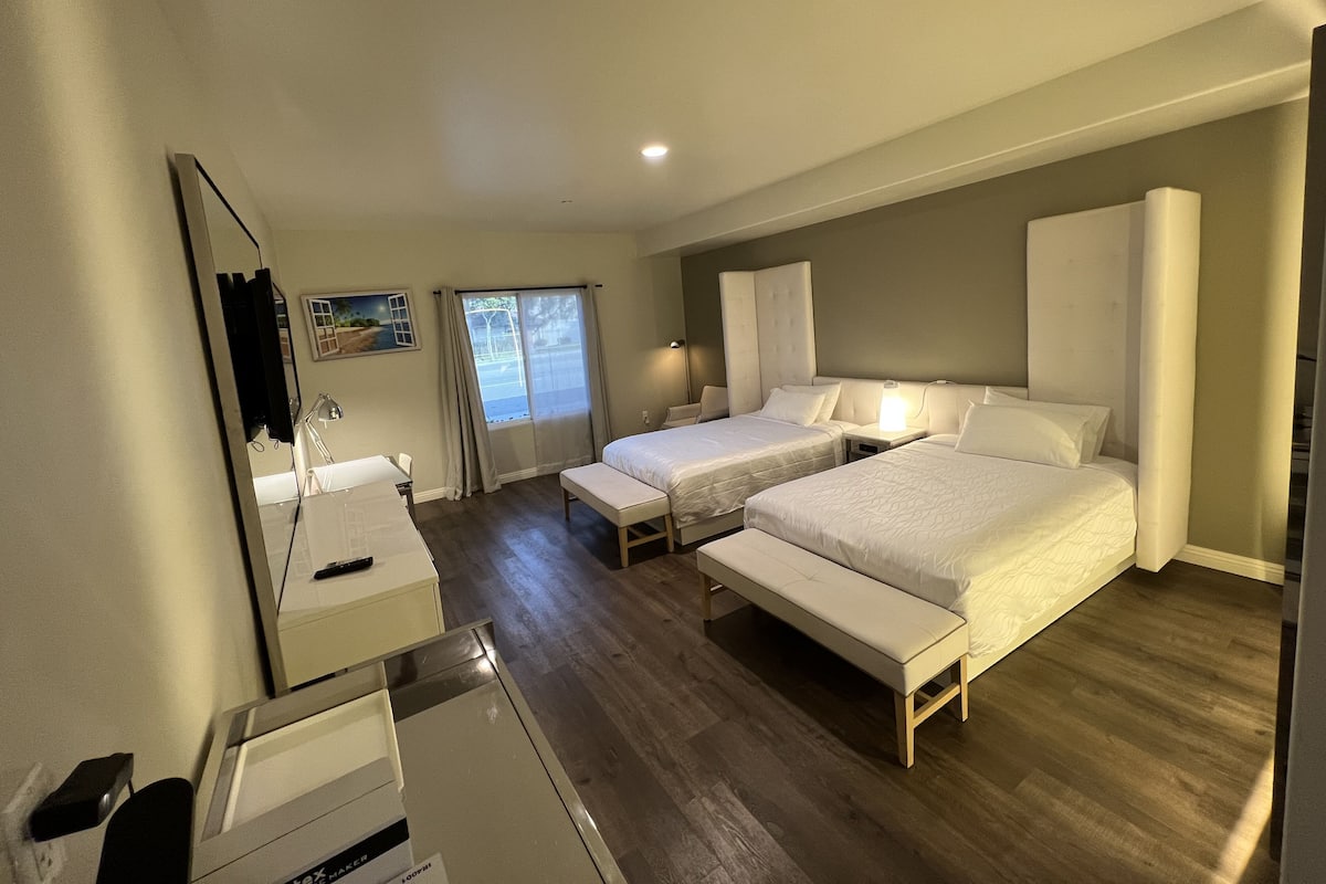 A hotel room with two beds, a bench, a desk, a mirror, and a window with curtains.