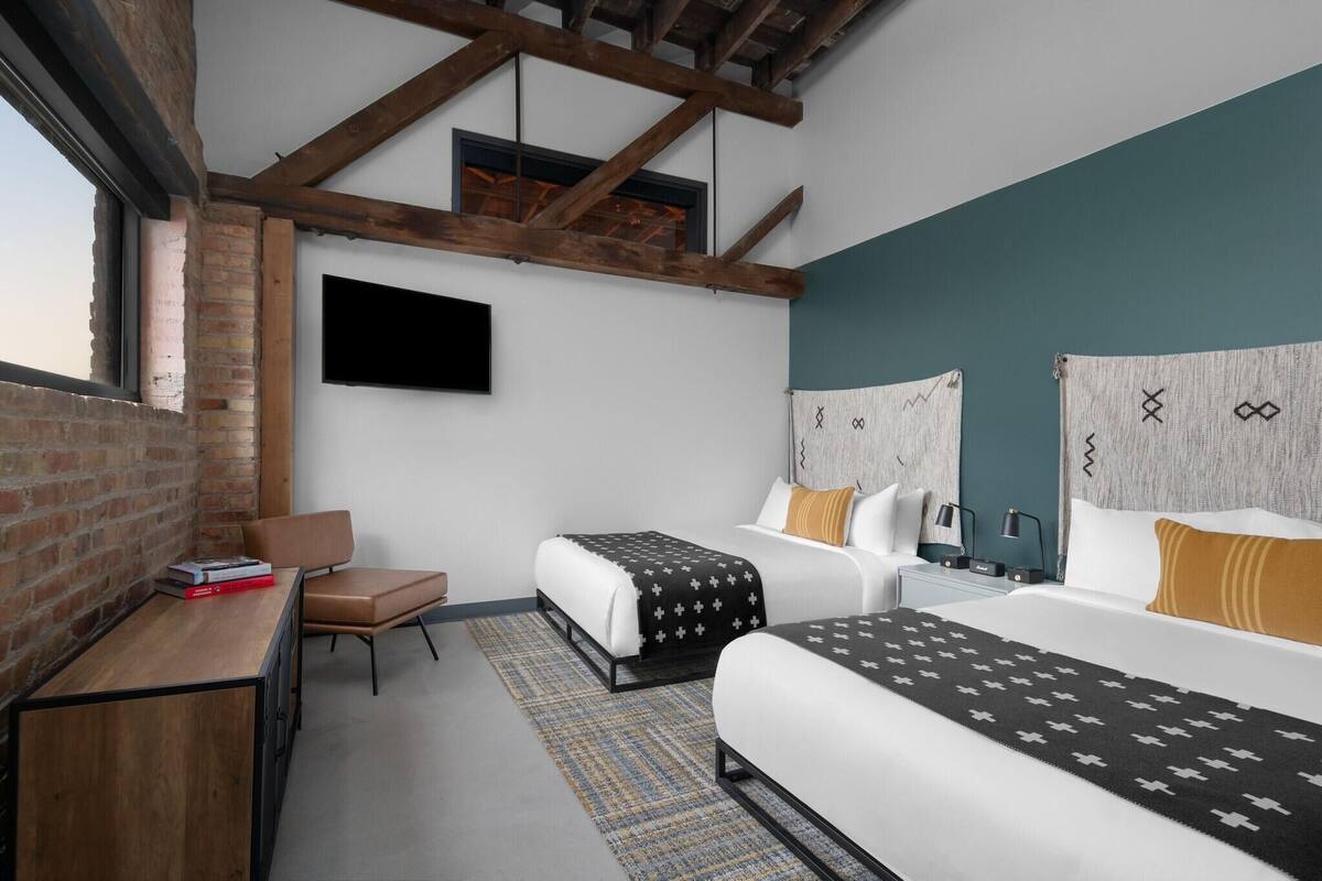 A modern hotel room with exposed brick walls, a loft-style ceiling, two beds with patterned bedding, a desk with a chair, and a flat-screen TV.