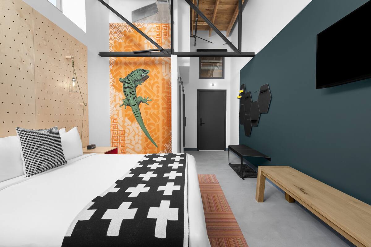 A modern hotel room with a large bed, a wooden headboard, a wall-mounted TV, and a unique green dinosaur wall decoration.