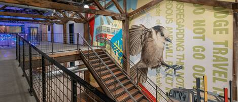 Una escalera con barandillas de metal que conduce a un piso superior, vigas de madera en el techo y un mural con un pájaro y un tren.