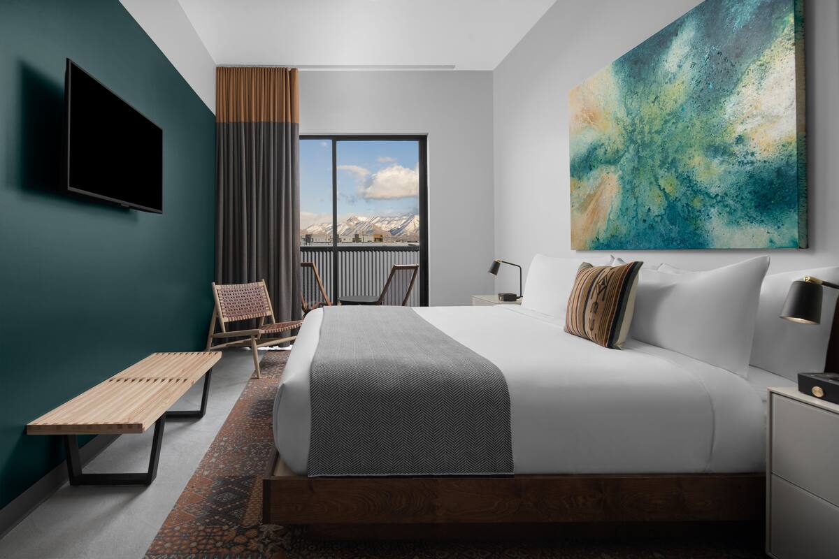 A modern hotel room with a large bed, a TV mounted on the wall, a balcony with a view of mountains, and a wooden bench.
