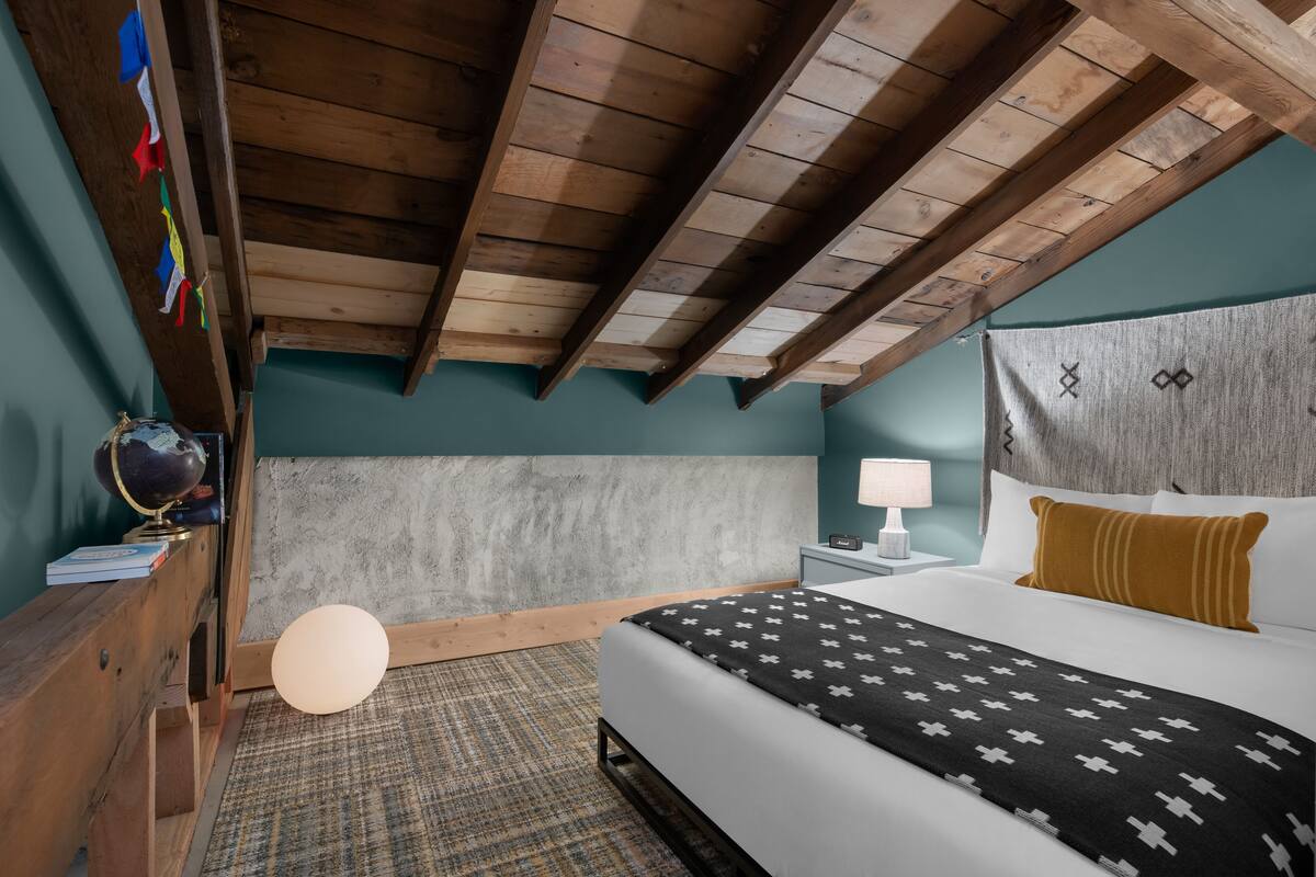 A bedroom with a wooden ceiling, a bed with a patterned blanket, a nightstand with a lamp, and a globe on a shelf.