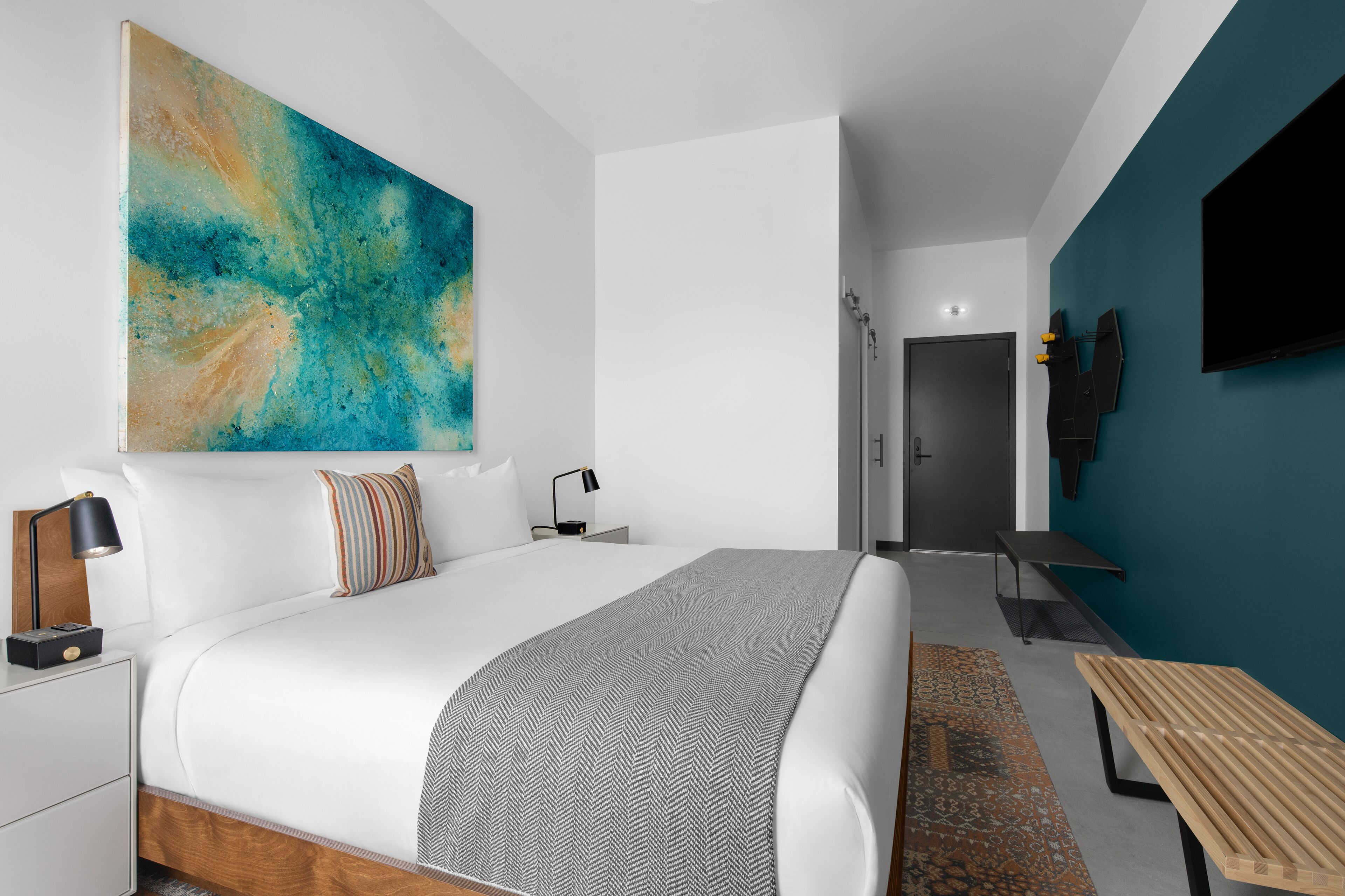 A modern bedroom with a large bed, a bedside table, a bench, a TV mounted on the wall, and a door leading to a bathroom.