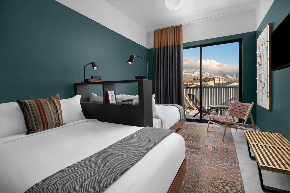 A hotel room with a large bed, a desk with a computer, a chair, and a view of the mountains through the window.