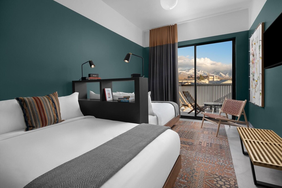 Quarto de hotel com uma cama grande, uma escrivaninha com computador, uma cadeira e vista para as montanhas pela janela.