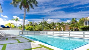 A rectangular swimming pool with a white deck and green grass edging, surrounded by palm trees and a clear blue sky. - Luxury 5BR/3BA Villa Walk to  Hollywood Beach (Hollywood)